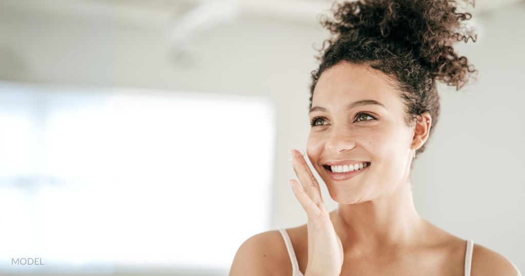 Beautiful woman with glowing clear skin is smiling while holding here face. (Model)