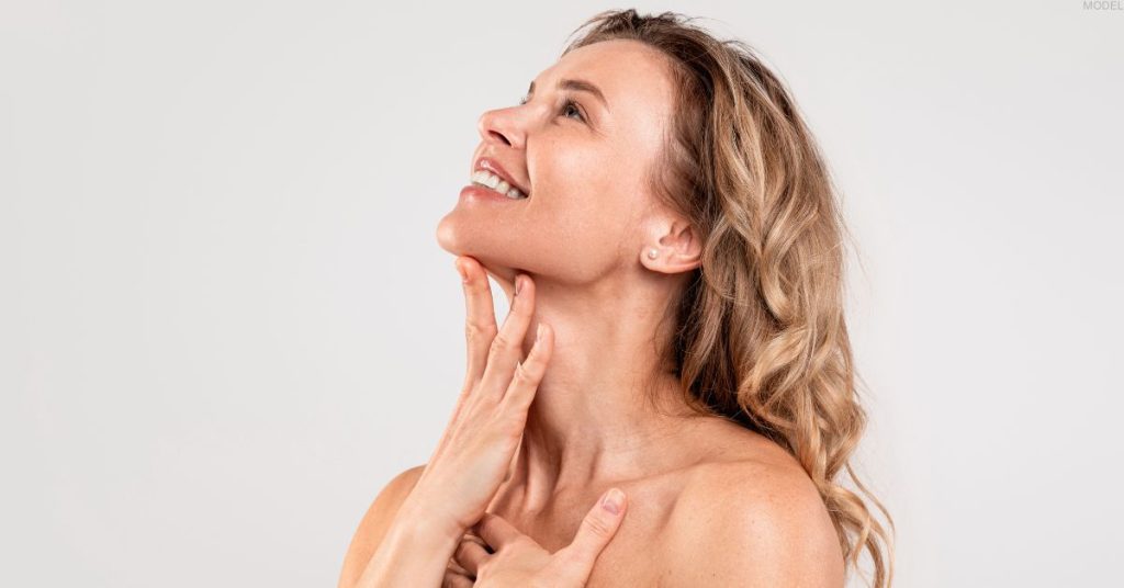 A woman smiles, looking upwards, with her hand gently touching her neck. (MODEL)