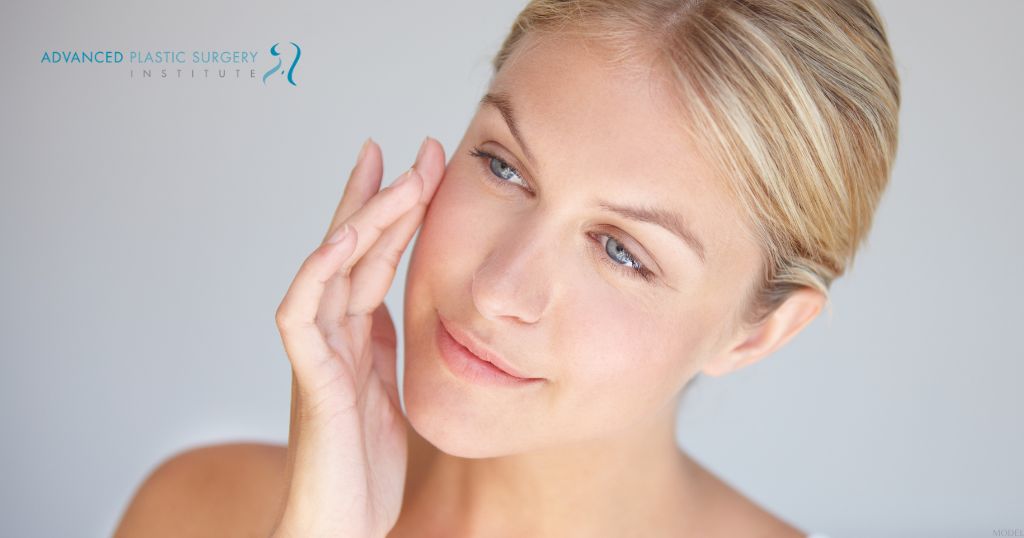 a woman touching her face and looking at the camera (MODEL) Advanced Plastic Surgery Institute Logo