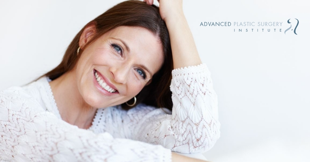 Middle age woman is smiling while sitting on a couch (model) Advanced Plastic Surgery Institute Logo